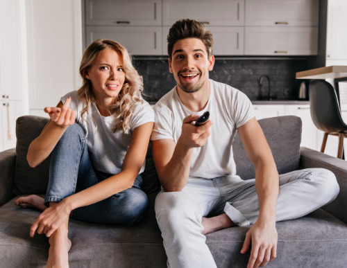 TV ou projetor: qual é a melhor escolha para o entretenimento? 1 Homem segurando controle da TV com expressão sorridente e mulher ao seu lado com expressão de dúvida.